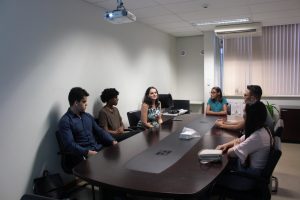 Em uma sala do TJMMG estão sentados em volta de uma mesa de reunião seis pessoas. A terceira da esquerda para a direita é a servidora Cynthia Chiari Barros, da Diretoria de Recursos Humanos. Ela é branca, tem cabelos castanhos e usa uma blusa estampada. Ela fala com as outras cinco pessoas, todas jovens, sendo três homens e duas mulheres aprovados na seleção para provimento de vagas de estágio na Justiça Militar do estado de Minas Gerais