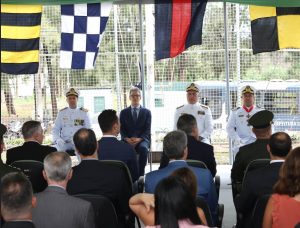 A imagem mostra momento da passagem de comando da Capitania Fluvial de Minas Gerais. Há diversos convidados sentados na plateia, olhando para o dispositivo de honra, onde constam quatro homens sentados. O primeiro à esquerda da imagem é o capitão de mar e guerra Alessandro de Paula Lima, que veste uniforme branco da Marinha do Brasil. Ao lado dele está o governador Romeu Zema, de terno azul e óculos. Após o governador há outros dois oficiais da Marinha, sendo que o mais à direita é o capitão Leonardo Carvalho de Lucena Navaes, que deixa o posto de comando. Ele também veste o uniforme branco da Marinha.