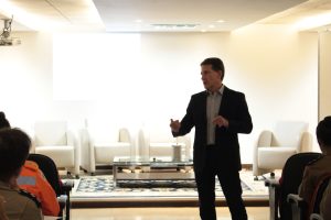 A imagem mostra o desembargador Fernando Antônio Nogueira Galvão da Rocha falando em pé no auditório dop TJMMG. Ele veste terno escuro e camisa clara. Ao fundo há poltronas brancas vazias, uma mesa de centro sobre um tapete. Em primeiro plano há detalhes da plateia, formada por pessoas com a farda da PMMG e do CBMMG