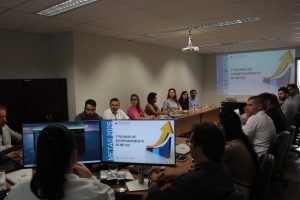 Imagem mostra uma sala de reunião com pessoas, entre homens e mulheres, sentadas diante de uma mesa de madeira. Em primeiro plano está uma tela de computador com uma seta amarela e o texto "Metas 2025 - 1ª reunião de acompanhamento de metas". A mesma imagem é projetada na parede