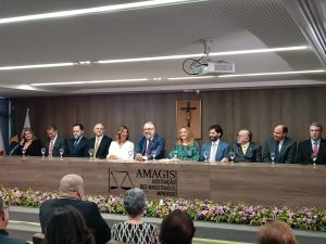 A imagem mostra uma mesa de autoridades durante a solenidade. Onze pessoas, sendo três mulheres e oito homens, estão sentados atrás de uma bancada de madeira, que tem o logotipo da Amagis gravado na frente. Eles usam trajes formais e exibem expressões sorridentes. O fundo tem um painel de madeira com um crucifixo centralizado. Acima da mesa, no teto branco, há um projetor. No primeiro plano, algumas pessoas do público aparecem de costas. Na parte inferior da imagem, há um arranjo de flores coloridas decorando a frente da bancada.