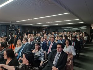 A imagem mostra a plateia sentada no auditório, entre homens e mulheres presentes. A juíza Renata Rodrigues de Pádua, de blusa branca e saia marrom com estampa, sorri para a imagem. Ao seu lado está o juiz João Pedro Hoffert Monteiro de Lima, de terno escuro, camisa branca e gravata rosa, e o juiz George Walter Barreto Paviotti, de camisa branca e gravata azul