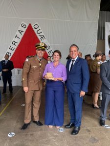 Há três pessoas no pátio onde foi realizada a entrega da medalha de Mérito da Inteligência. Ao fundo há a bandeira de Minas Gerais. À esquerda está o diretor de Inteligência da PMMG, que veste a farda da Polícia Militar. Ao centro está a sargento agraciada com a moeda comemorativa. Ela está de cabelos presos, veste camisa lilás, calça azul e segura uma caixa com a homenagem recebida. À direita está o vice-presidente do TJMMG, que veste terno azul. Os três sorriem para a foto.