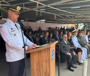 Na imagem está o coronel Erlon Dias do Nascimento Botelho em pé, falando ao microfone, em um púlpito de madeira. Ele veste farda branca, chapéu e calças pretas. Há pessoas sentadas ao fundo da fotografia, incluindo o desembargador Jadir Silva, que está situado na primeira fileira de autoridades. O desembargador veste terno cinza com gravata amarela e usa óculos