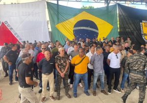 Na imagem está um grupo de homens em pé, e ao fundo há três bandeiras. À esquerda está a bandeira de Minas Gerais; ao meio, a do Brasil; e à direita, a da Rotam