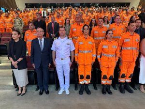 A imagem mostra a plateia durante a solenidade de posse do novo chefe do Estado-Maior do CBMMG, em sua maioria formada por integrantes do Corpo de Bombeiros. Todos estão de pé. Na primeira fileira, há seis pessoas, entre homens e mulheres. O segundo da esquerda para direita é o desembargador coronel Osmar Duarte Marcelino, que usa terno azul escuro, gravata azul e óculos. Ao seu lado direito está o capitão dos Portos de Minas Gerais, que veste farda branca da Marinha, e os coronéis Daniela Lopes, Jordana Daldegan e Moisés Magalhães de Sousa, que vestem farda laranja do CBMMG