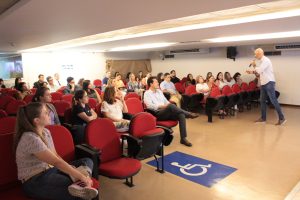 A imagem mostra o auditório do TJMMG com cadeiras vermelhas, onde estão sentados servidores e colaboradores voluntários do projeto, entre homens e mulheres. Eles olham o servidor que conduz a oficina de camisa listrada e calça jeans