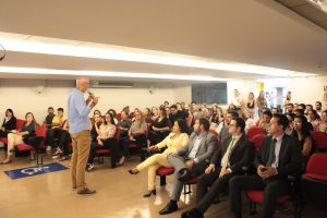 A imagem mostra o servidor do TJMMG Gustavo Cândido da Silva vestindo camisa social azul e calça marrom. Ele discursa para várias pessoas. O auditório tem paredes brancas, iluminação clara e cadeiras vermelhas dos dois lados, onde os convidados estão sentados