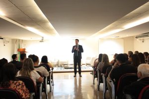 A foto mostra o desembargador Fernando Armando Ribeiro vestindo terno cinza e gravata vermelha. Ele está em pé no centro do auditório do TJMMG discursando e há pessoas sentadas dos dois lados da sala. As paredes são brancas e a iluminação é clara