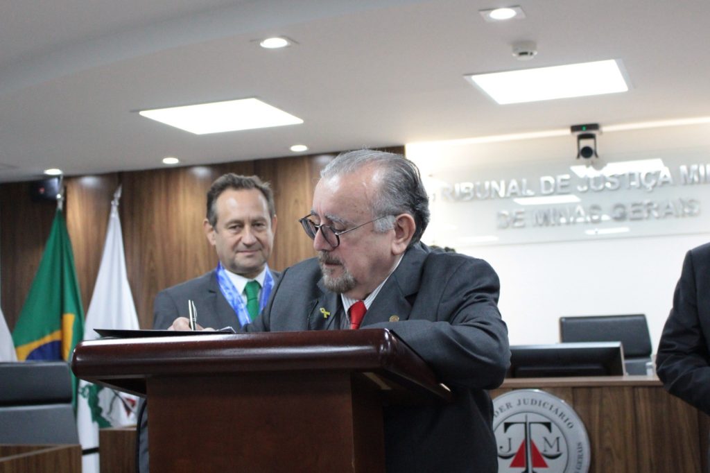 As fotos representam o momento de assinatura de cada um dos quatro desembargadores na sala do Tribunal Pleno do TJMMG. Todos estão de terno e assinaram em um púlpito de madeira