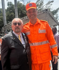 A foto registra duas pessoas lado a lado em uma área aberta. À esquerda está o desembargador Jadir Silva vestindo terno cinza com gravata amarela e usando óculos. À direita está o coronel Moisés Magalhães de Sousa, com farda laranja dos bombeiros.