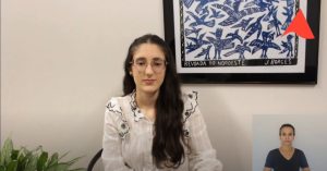 A imagem mostra trecho do vídeo apresentado pela oficial judiciário do TJMMG Juliana Bueno Lima de Aguiar. Ela usa camisa branca com detalhes pretos e óculos. O ambiente está decorado com uma planta e um quadro de xilogravura com representação de pássaros azuis. No canto superior direito há um detalhe da logomarca do TJMMG e no canto inferior há um intérprete de libras vestindo camisa preta.