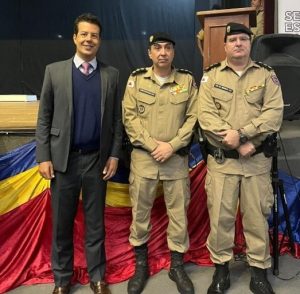 Imagem mostra o desembargador Fernando José Armando Ribeiro ao lado do coronel Cintra e do tenente coronel Henrique. Eles estão em pé, na frente do palco de um auditório que tem um pano nas cores vermelho, azul e amarelo. O desembargador está à esquerda, usando um terno com camisa azul clara, gravata vermelha e suéter azul, enquanto os militares usam a farda da PMMG