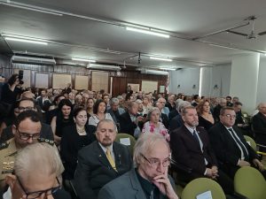A foto mostra o auditório do IHGMG com diversos convidados da solenidade de posse da presidência do IHGMG sentados na plateia, entre homens e mulheres. O presidente do TJMMG está sentado na segunda fileira, vestindo terno preto e gravata amarela