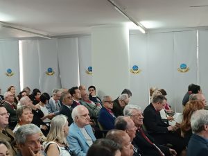 A foto mostra o auditório do IHGMG com diversos convidados da solenidade sentados na plateia, entre homens e mulheres. Ao fundo da imagem há uma cortina branca com a logomarca da entidade, um brasão em azul e amarelo