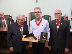 A imagem mostra em primeiro plano três pessoas, sendo que ao meio está o novo presidente de honra do IHGMG, governador Romeu Zema, usando camisa social azul, calça preta e óculos. Ele segura em sua mão direita um troféu com um almocafre, uma antiga ferramenta utilizada na mineração; em sua mão esquerda, o diploma de honra da Casa João Pinheiro; e em seu pescoço, está a medalha João Pinheiro, com uma fita vermelha. As outras duas pessoas ao lado, bem como as sentadas ao fundo em uma mesa solene de madeira com cadeiras verdes, vestem ternos com gravatas coloridas, usam óculos e também estão com a medalha João Pinheiro