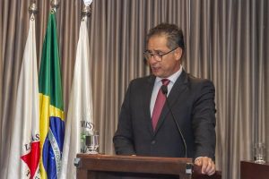 A imagem mostra o novo ouvidor no MPMG, promotor de Justiça Rolando Carabolante, usando terno, gravata vermelha e óculos. Ele fala ao microfone em um púlpito de madeira. Ao fundo há uma cortina dourada e à esquerda há três bandeiras: uma do Estado de Minas Gerais, uma do Brasil e a bandeira do Ministério Público