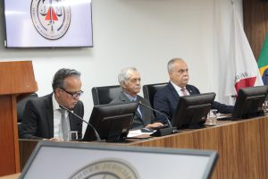 Na foto estão três desembargadores do TJMMG sentados de frente para o computador. O primeiro é o vice-presidente James Ferreira Santos vestindo terno preto e gravata cinza; o segundo é o corregedor Sócrates Edgard dos Anjos, com terno e gravata cinza; e o terceiro é Rúbio Paulino Coelho, com terno azul escuro e gravata vermelha. Ao fundo uma tela projeta o brasão do TJMMG e no canto direito estão posicionadas as bandeiras de Minas Gerais e do Brasil