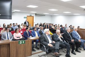 A imagem mostra várias pessoas sentadas na plateia da sala do Tribunal Pleno do TJMMG. Na primeira fileira, à esquerda, está o ex-procurador de Justiça Epaminondas Fulgêncio Neto usando terno cinza, gravata azul e óculos. Ao seu lado estão três ex-presidentes do Tribunal: Décio de Carvalho Mitre, com terno preto e gravata listrada; seguido por Paulo Duarte Pereira, com terno preto e gravata cinza; e por último, Luís Marcelo Inacarato, com terno azul e gravata branca