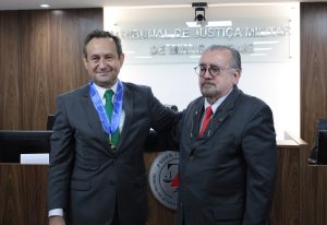 A foto mostra dois desembargadores lado a lado na sala do Tribunal Pleno do TJMMG. À esquerda está o presidente do TJMG Luiz Carlos de Azevedo Corrêa Junior, vestindo terno cinza e gravata verde, e à direita o presidente do TJMMG Jadir Silva, com terno preto, gravata vermelha e óculos. Ao fundo há um letreiro com o nome do TJMMG