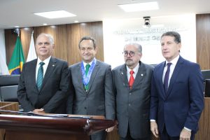 A imagem mostra quatro desembargadores na sala do Tribunal Pleno do TJMMG. À esquerda está o segundo vice-presidente do TJMG Saulo Versiani Penna, trajando um terno preto com gravata azul, seguido pelo presidente do TJMG Luiz Carlos de Azevedo Corrêa Junior, com terno cinza e gravata verde. A terceira pessoa é o presidente do TJMMG Jadir Silva, vestindo terno cinza e gravata vermelha, e por último está o diretor da EJM Fernando Antônio Nogueira Galvão da Rocha, com terno azul e gravata roxa