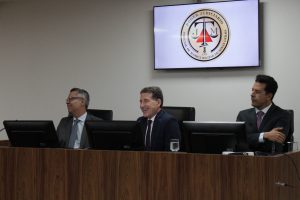 A imagem mostra três desembargadores do TJMMG sentados de frente para o computador. O primeiro é Osmar Duarte Marcelino vestindo terno preto e gravata cinza; o segundo é o diretor da EJM Fernando Antônio Nogueira Galvão da Rocha, com terno azul escuro e gravata roxa; e o terceiro é o ouvidor Fernando Armando Ribeiro, com terno preto e gravata cinza. Ao fundo uma tela projeta o brasão do TJMMG