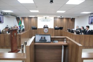 A imagem mostra oito homens sentados na sala de julgamento do Tribunal Pleno, todos em tribunas de madeira dispostas com microfones e telas de computador. Três são desembargadores que estão à esquerda da imagem, vestindo trajes militares, e três outros desembargadores estão à direita, sendo um com traje militar e outros dois com a beca de magistrados civis. Ao centro está o presidente do TJMMG, Jadir Silva, e um servidor, ambos vestidos com beca. Ao fundo está escrito Tribunal de Justiça Militar de Minas Gerais, e na frente o brasão do órgão. Em primeiro plano, há um computador exibindo a transmissão ao vivo do evento, focada na imagem do presidente.