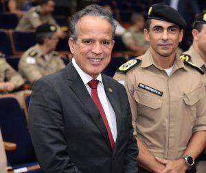 A imagem mostra o desembargador coronel James Ferreira Santos vestindo terno preto, camisa branca e gravata vermelha. Ao seu lado direito está o comandante-geral da PMMG com farda marrom e boina. Eles estão na plateia de um auditório, e ao fundo estão outros policiais militares sentados, todos vestindo farda marrom