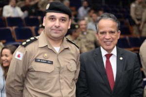 A foto mostra o novo comandante da Terceira Região da PMMG vestindo farda marrom e boina. Ao seu lado direito está o desembargador coronel James Ferreira Santos vestindo terno preto, camisa branca e gravata vermelha. Eles estão na plateia de um auditório, e ao fundo estão outros convidados civis e policiais militares sentados vestindo farda marrom