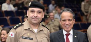 A foto mostra o novo comandante da Terceira Região da PMMG vestindo farda marrom e boina. Ao seu lado direito está o desembargador coronel James Ferreira Santos vestindo terno preto, camisa branca e gravata vermelha. Eles estão na plateia de um auditório, e ao fundo estão outros convidados civis e policiais militares sentados vestindo farda marrom