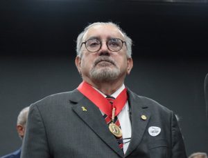 Imagem mostra um retrato fechado no busto do presidente do TJMMG, desembargador Jadir Silva. Ele veste terno preto, camisa branca, gravata listrada. Ele usa óculos, cavanhaque grisalho e usa um colar condecorativo com faixa vermelha