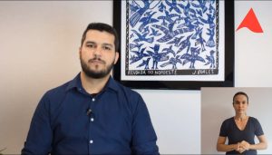 A imagem mostra o servidor Lucas Alves Edmundo Gomes sentado, vestindo camisa social azul. Ele tem cabelo e barba pretos. Ao fundo há um quadro de xilogravura com a representação de pássaros azuis. No canto superior direito há um detalhe da logomarca do TJMMG, e no canto inferior direito há uma intérprete de libras que está de cabelos presos e veste camiseta preta