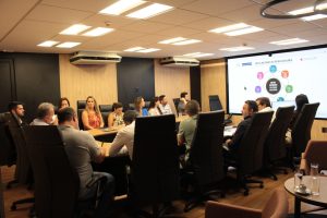 A imagem mostra uma sala de reunião com uma mesa reunindo homens e mulheres. O ambiente é composto por móveis e painel de parede em tons de madeira e preto. O grupo olha para uma tela de LED onde é projetado o novo layout do Painel de Metas Nacionais do Poder Judiciário, composto por ícones roxo, rosa, azul, laranja, verde e preto