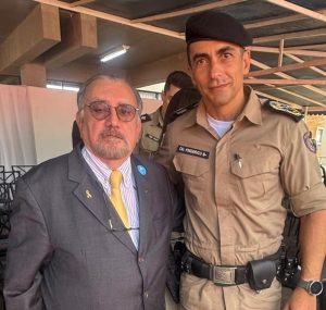 A foto registra duas pessoas lado a lado em um palanque. À esquerda está o desembargador Jadir Silva, vestindo terno cinza com gravata amarela e usando óculos. À direita está o comandante-geral da PMMG com farda marrom