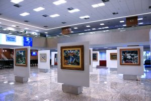 A foto mostra o hall do edifício-sede do TJMG, um espaço amplo e iluminado, com piso de mármore claro refletindo a luz do teto. Várias telas do artista Inimá de Paula estão expostas em painéis brancos fixados sobre bases de concreto, distribuídas pelo salão. As pinturas possuem molduras variadas, algumas em madeira lisa e outras ornamentadas, e retratam paisagens e cenas urbanas coloridas. Ao fundo, há uma parede de vidro com persianas abertas, permitindo a entrada de luz natural