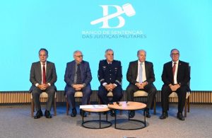 A foto mostra cinco homens, sendo quatro civis de terno e um militar fardado, sentados em cadeiras dispostas em um palco. O vice-presidente do TJMMG é o primeiro da esquerda para a direita. À frente deles há uma mesa de centro, e ao fundo um telão com a logomar com Banco de Sentenças formado pela letra B estilizada com um malhete, nome dado ao martelo que simboliza a Justiça