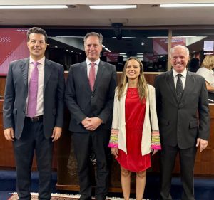A imagem mostra quatro pessoas em pé. À esquerda está o ouvidor do TJMMG, desembargador Fernando José Armando Ribeiro, vestindo terno azul escuro com gravata roxa. Ao seu lado está o novo ouvidor no MPMG, o promotor de Justiça Rolando Carabolante, usando terno azul e gravata vermelha. Ao lado deles há uma mulher de vestido vermelho e blaser branco, com destalhes coloridos nas mangas, e um homem de terno