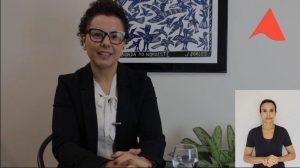 Imagem mostra a defensora pública sentada em frente a uma parede branca onde há um quadro branco e azul, e um vaso de plantas. Ela tem cabelos castanhos, usa óculos, e veste blazer preto e blusa branca. No canto direito inferior da imagem está a janela da intérprete de libras, uma mulher de camisa preta. No canto direito superior está a logomarca reduzida do TJMMG, formado pelo triângulo da bandeira de Minas Gerais estilizado.