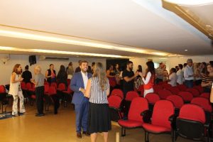 A imagem mostra o auditório do TJMMG com várias cadeiras vermelhas. O público, entre homens e mulheres, está em pé, dividido em duplas. No centro da imagem está o juiz João Pedro Hoffert Monteiro de Lima, de terno azul e camisa branca, acompanhado de uma servidora de blusa listrada e saia preta