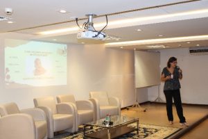 A imagem mostra uma mulher com cabelos castanhos e óculos, vestindo blusa estampada e calça preta, ela segura um microfone enquanto apresenta um slide projetado na parede. No ambiente, há quatro poltronas bege, uma mesa de vidro com microfone e copos, além de um quadro branco ao fundo.