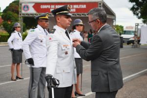 A foto mostra bombeiros com farda branca e o desembargador do TJMMG de terno cinza. Ele coloca uma medalha no aspirante Rodolfo Finamor Costa Gomes, que segura uma espada