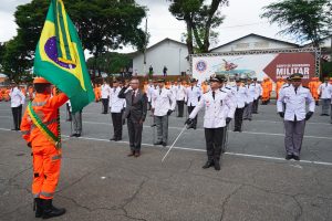 A imagem mostra momento da solenidade de formatura em que a turma de formandos, vestidos com a farda branca do CBMMG, está no pátio da Academia de Bombeiros Militar diante de um militar de farda laranja carregando a bandeira do Brasil. O desembargador do TJMMG está na frente da turma, prestando continência à bandeira