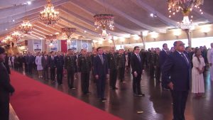 A imagem mostra homenageados na solenidade de comemoração aos 217 anos da JMU, entre homens e mulheres. Todos estão em um amplo salão, de pé em posição de respeito, e olham para frente. No chão há um longo tapete vermelho e no teto há vários lustres