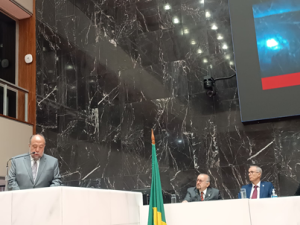 A imagem mostra o Plenário Juscelino Kubitschek da ALMG, um ambiente com parede de granito escuro, bancada e púlpito de mármore branco. Há três homens na imagem. O professor Hermes Vilchez Guerrero veste um terno cinza e gravata listrada. Ele está de pé, falando ao microfone atrás do púlpito. Ao lado está sentado o presidente do TJMMG usando terno preto, gravata e óculos. Entre eles há uma bandeira do Brasil.