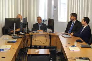 A imagem mostra quatro homens. O corregedor da Justiça Militar está ao centro, vestindo terno e gravata cinza, e ao seu lado estão os juízes Marcos Luiz Nery Filho e George Walter Barreto Paviotti, ambos vestindo terno escuro e gravata azul. Eles estão sentados em uma mesa de reunião com telas e notebook. Ao fundo há uma parede branca com janelas amplas e uma cortina clara