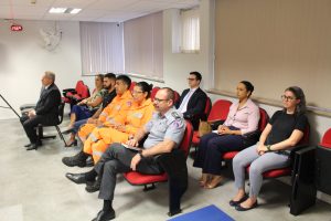 A imagem mostra homens e mulheres, civis e militares, sentados na plateia da sala de audiências da 1ª AJME. Na sala há cadeiras vermelhas, janelas amplas com persiana e as paredes são brancas