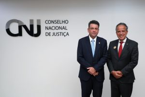 A imagem mostra dois homens em pé, diante de uma parede com o letreiro escrito CNJ Conselho Nacional de Justiça. Ambos estão de terno escuro e com as mãos cruzadas em frente ao corpo. O homem mais à direita da imagem é o vice-presidente do TJMMG, que tem cabelos grisalhos, gravata vermelha e sorri para a foto. Ao seu lado está o novo conselheiro do CNJ, de gravata azul