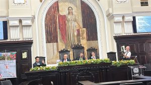 A imagem mostra quatro homens sentados em uma sala do Palácio da Justiça do Tribunal de Justiça de Pernambuco. Eles estão atrás de uma bancada de madeira ornada com flores, todos vestem terno e gravata. O desembargador do TJMMG está à esquerda da imagem, falando ao microfone. No lado oposto há um fotógrafo. A sala tem paredes em tom bege com uma grande ilustração com a deusa da Justiça