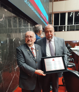 A imagem mostra o presidente do TJMMG, desembargador Jadir Silva, usando terno preto, gravata listrada e óculos. Ao lado está o professor Hermes Vilchez Guerrero vestindo terno cinza e gravata listrada. Os dois seguram a placa de alumínio com o título de Cidadão Honorário de Minas Gerais. Ao fundo da foto há uma parede branca com janelas e do lado esquerdo há uma parede de mármore preto e o telão do Plenário da ALMG