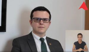 Imagem mostra um trecho do vídeo apresentado pelo juiz de Direito Bruno Cortez Torres Castelo Branco. Ele veste terno escuro, camisa branca, e está numa sala com parede branca e um detalhe de um quadro ao fundo